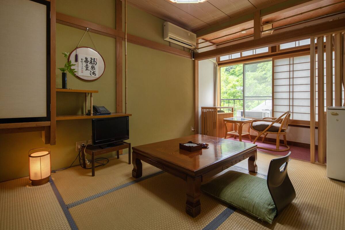 A traditional Japanese room with tatami flooring, a low wooden table, a television, and a sliding door to a balcony.