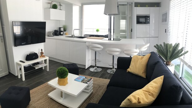 A modern living room with a black sofa, white coffee table, and a TV. There is a kitchen area with white cabinets and a dining space with bar stools. - Appt NEW Arcachon Center, Beach 250 m - Terrace - Private car park (Arcachon)