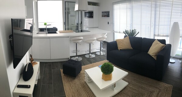 A modern living space with a kitchen, a black sofa with cushions, a white coffee table with a plant, and a remote control on a side table. - Appt NEW Arcachon Center, Beach 250 m - Terrace - Private car park (Arcachon)