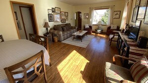 A spacious living room with a wooden floor, a sofa, a coffee table, and a TV. There are framed pictures on the wall and a window with curtains. - Prairie Cottages in historic Rapid City (Rapid City)