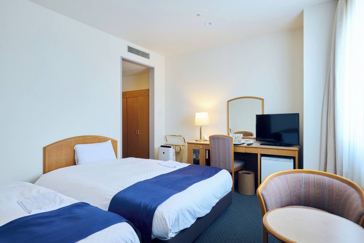 A hotel room with two beds, a desk with a television, a chair, and a lamp.