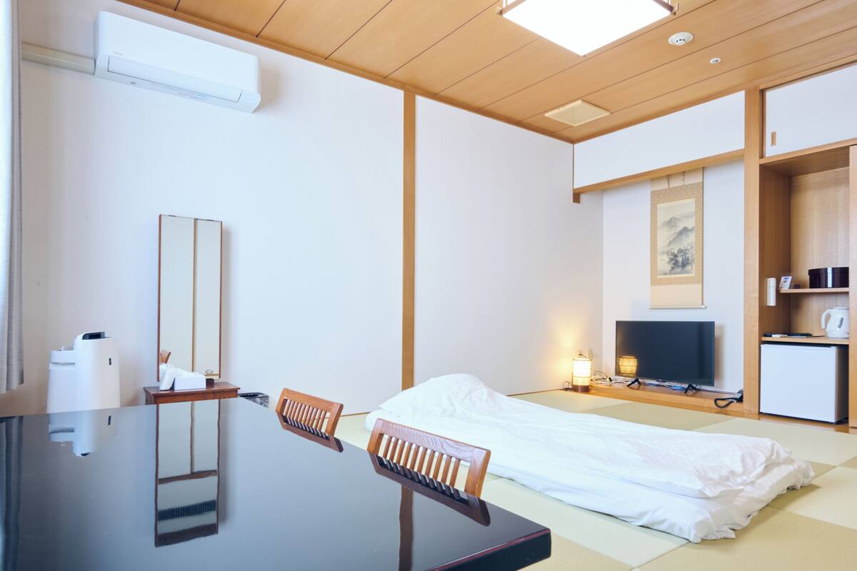 A hotel room with a bed, a desk with a chair, a television, a small refrigerator, and a wall-mounted air conditioner.