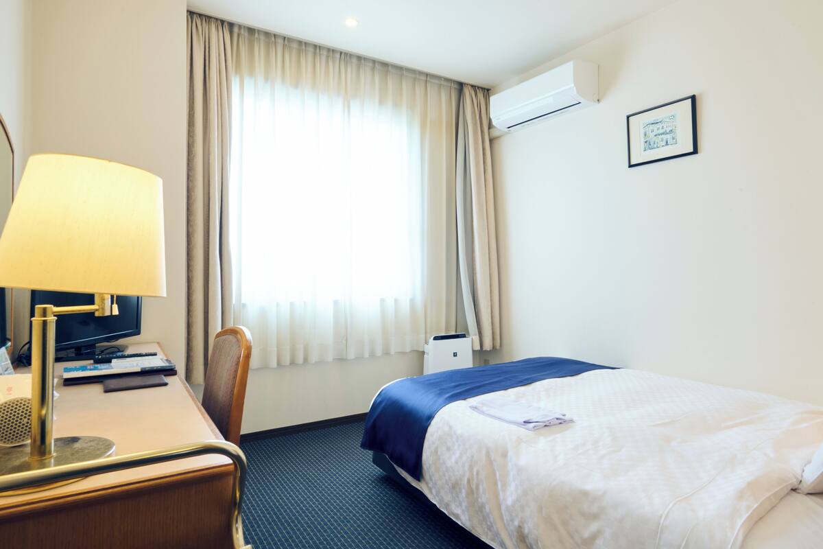 A hotel room with a bed, a desk with a lamp, a chair, a television, and a window with curtains.