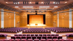 Un auditorio amplio con hileras de asientos morados, un escenario con un atril y varios proyectores instalados en el techo.