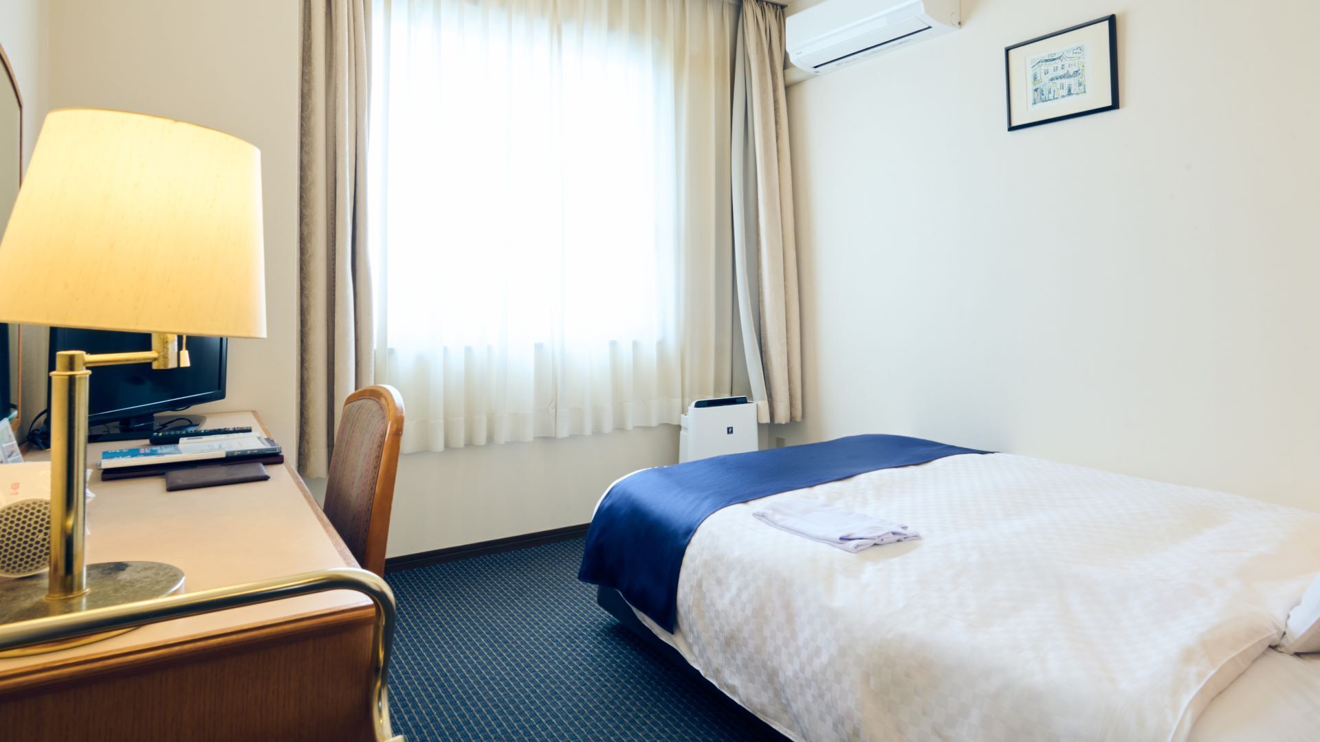 A hotel room with a bed, desk, chair, lamp, and a window with curtains.