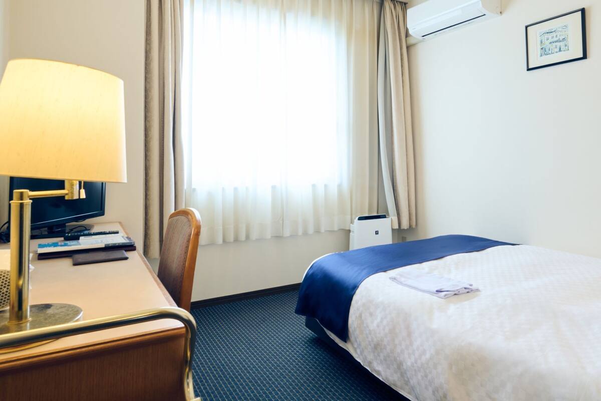 A hotel room with a bed, desk, chair, lamp, and a window with curtains.