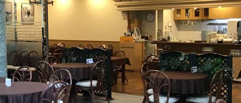 A dining area with round tables and wicker chairs, a kitchen in the background, and a chandelier above.