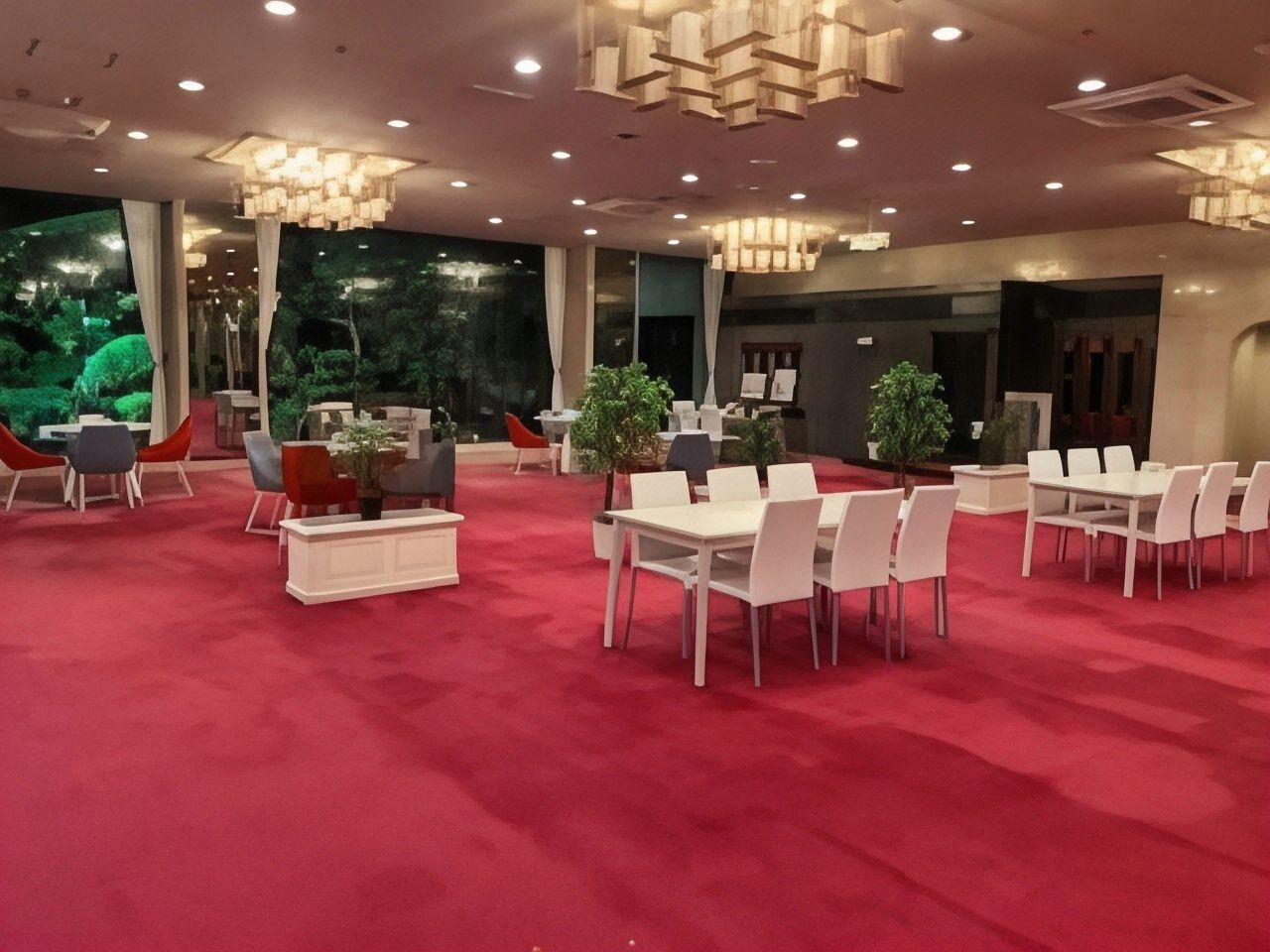 A spacious indoor area with multiple tables and chairs, a red carpet floor, and a large window with a view of greenery.