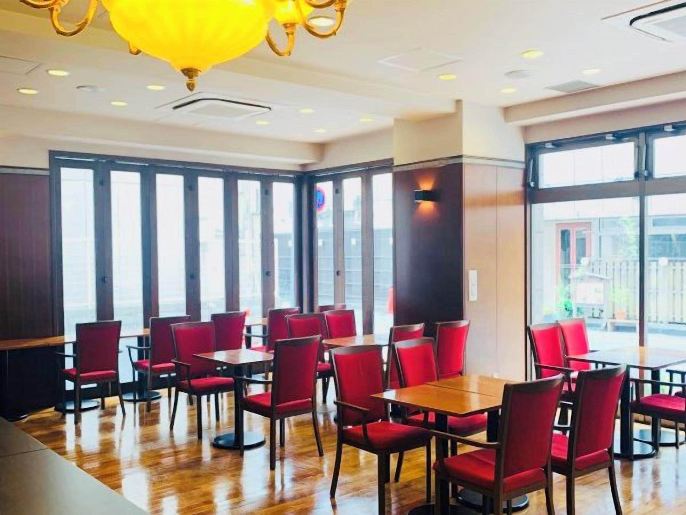 A restaurant with red chairs and tables, large windows, and a yellow chandelier.