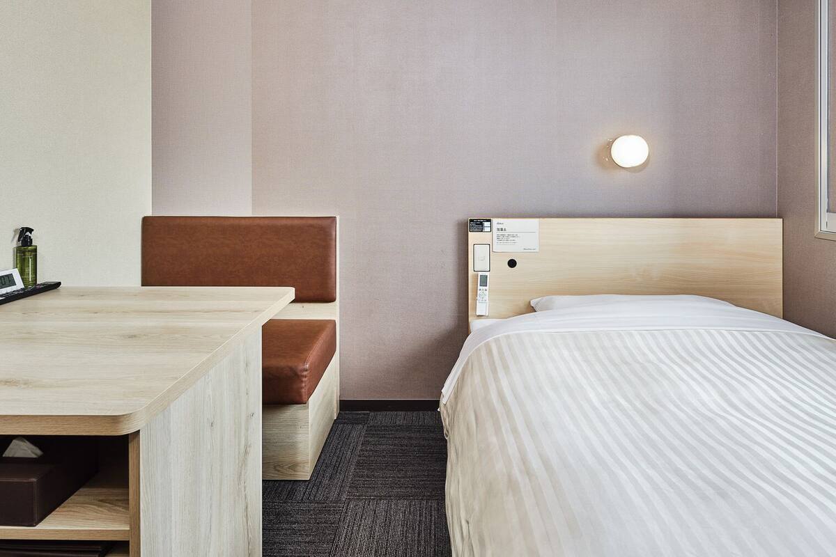 A hotel room with a wooden desk, a brown cushioned bench, a bed with a striped bedspread, a nightstand with a lamp, and a wall-mounted control panel.