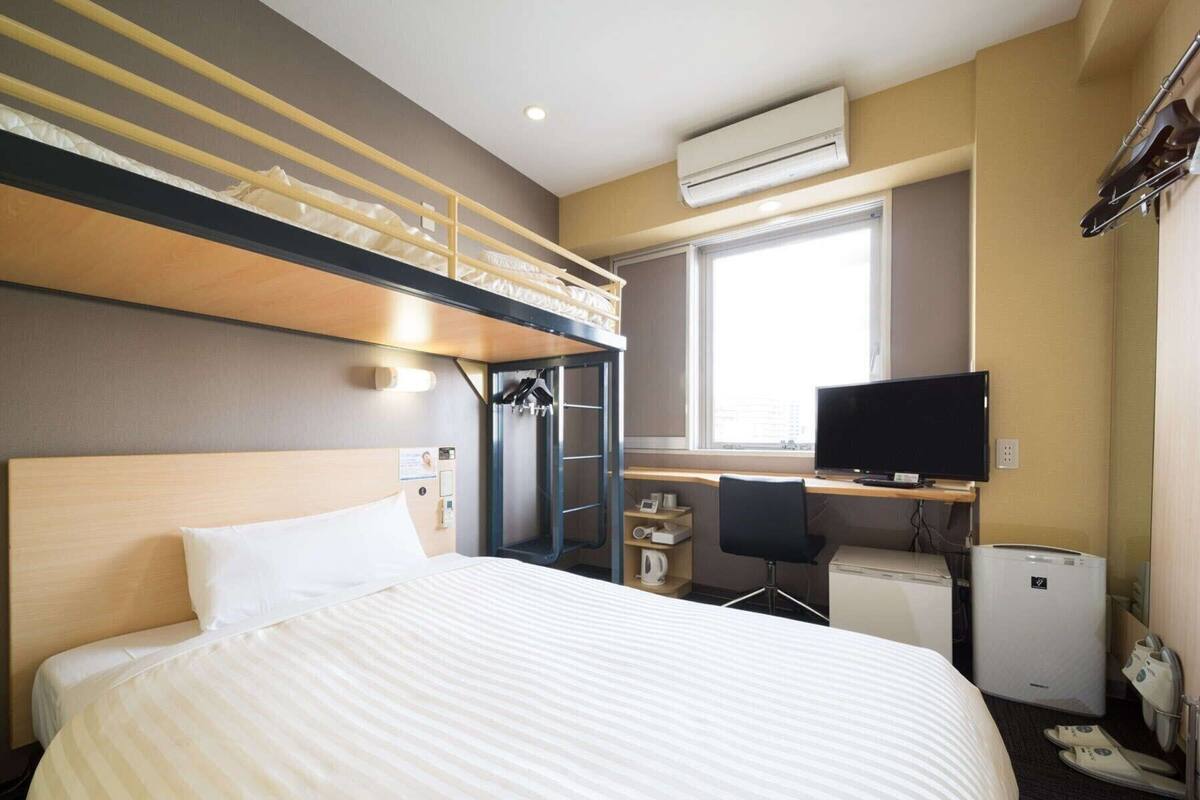 A hotel room with a bunk bed, a desk with a computer, a TV, and a small kitchenette.