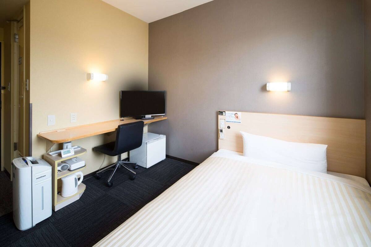 A hotel room with a bed, a desk with a computer, a chair, a small refrigerator, and a coffee maker.