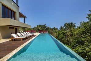 Un área de piscina moderna con sillones de descanso y una terraza de madera, rodeada de exuberante vegetación y palmeras.