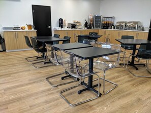 A modern dining area with clear chairs, black tables, and a kitchen counter with coffee machines and food displays.