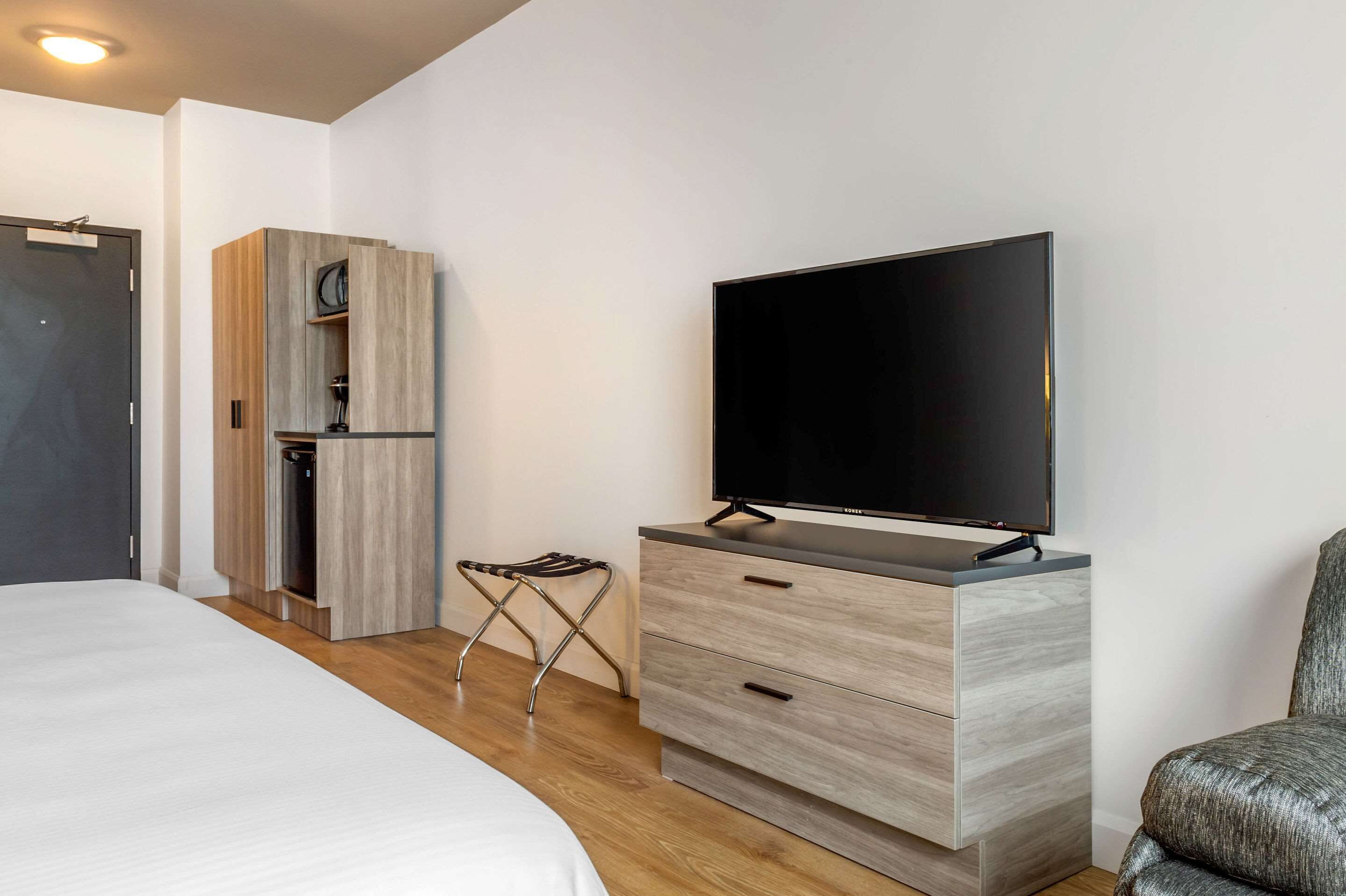 A modern hotel room with a bed, a TV on a wooden cabinet, a small kitchenette, and a chair.