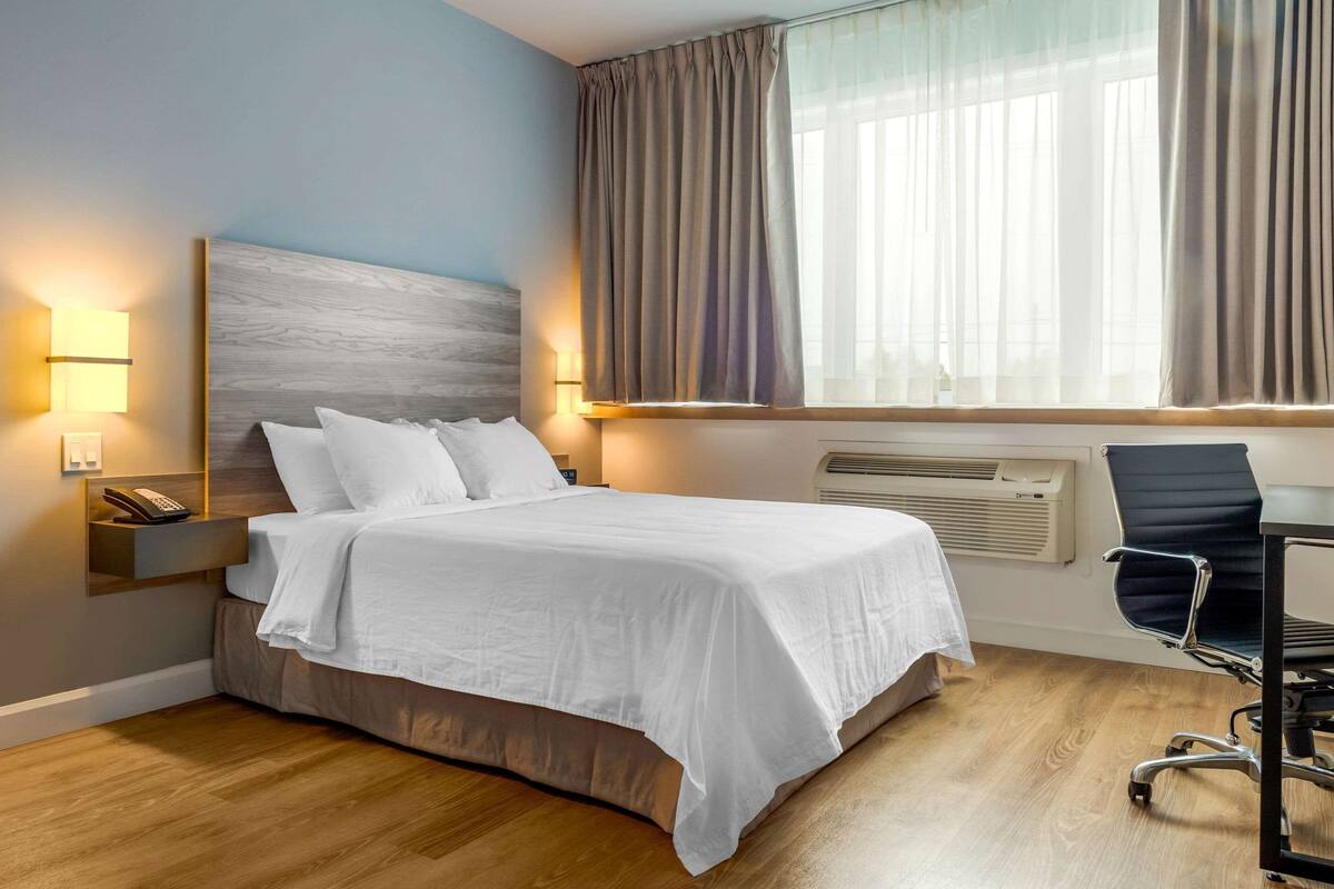 A hotel room with a bed, bedside lamp, air conditioning unit, and a desk with a chair.