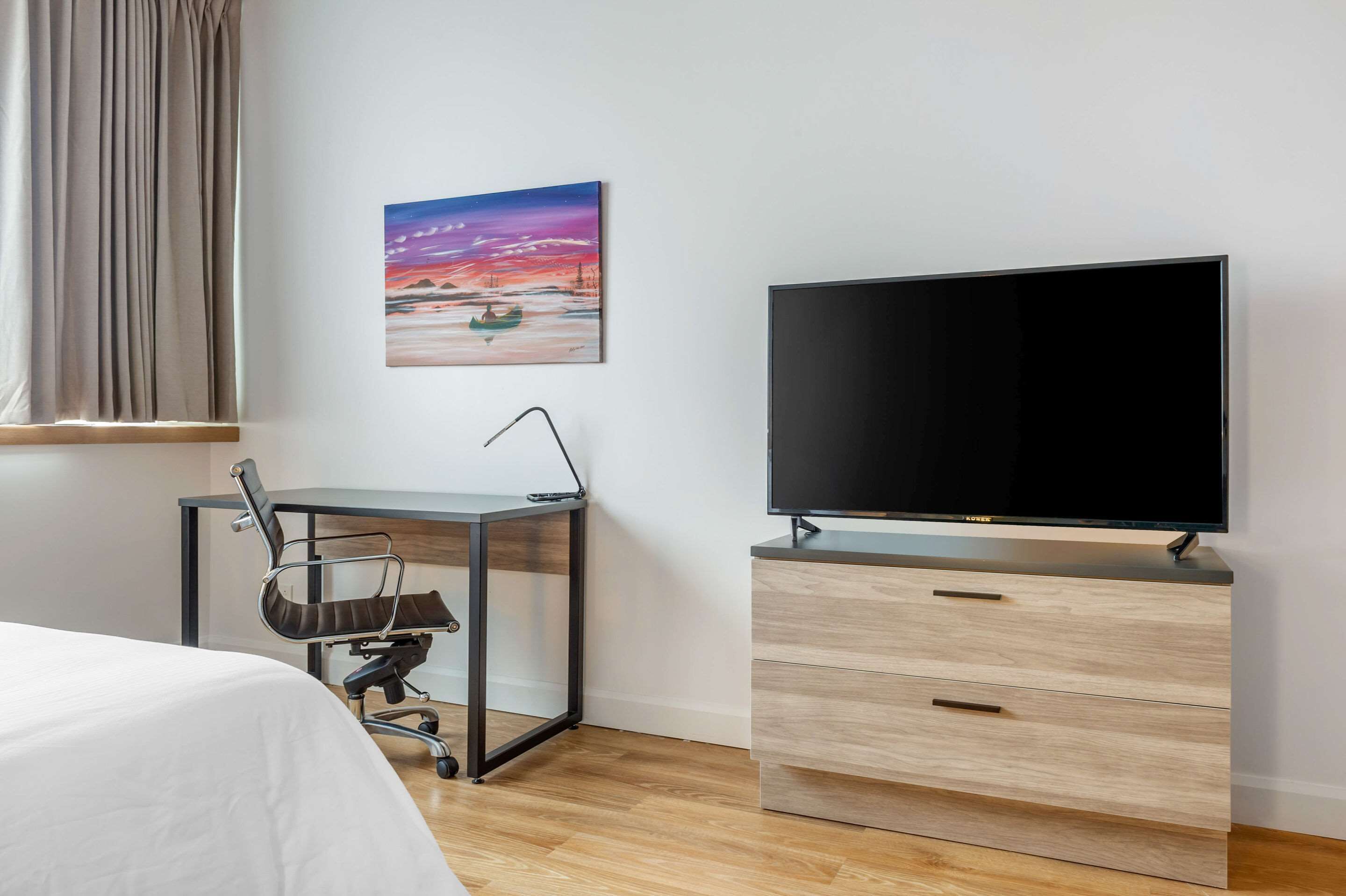 A hotel room with a bed, a desk with a chair, a TV on a wooden cabinet, and a painting on the wall.