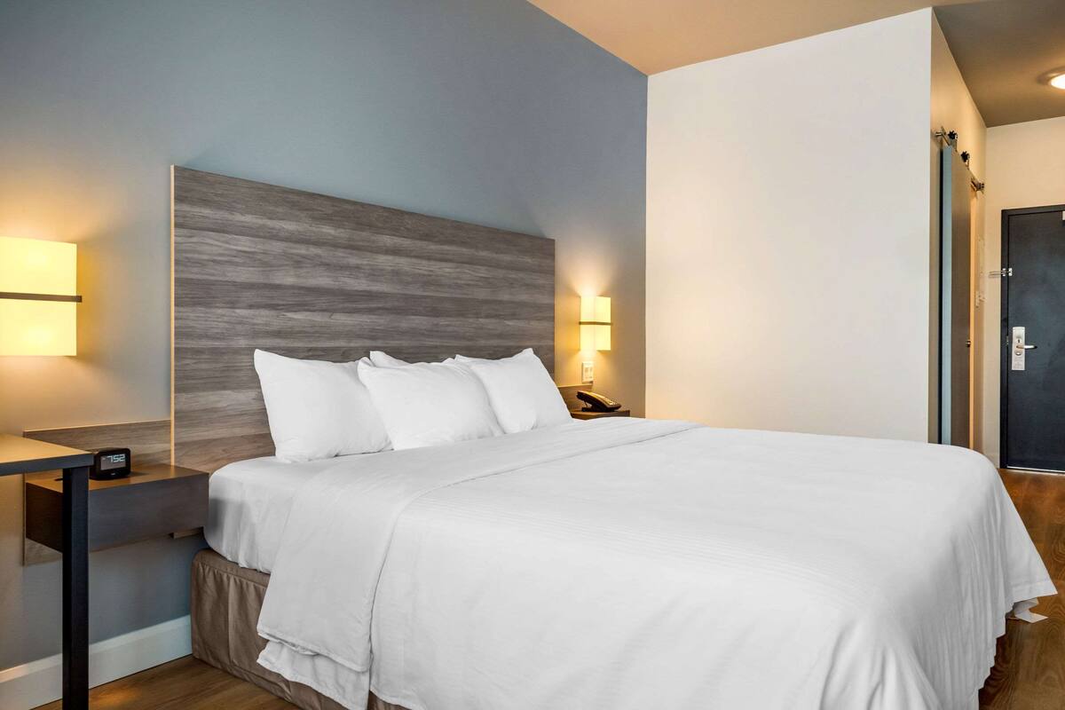 A neatly made bed with white linens in a modern room with a wooden headboard, a bedside table with a lamp, and a wall-mounted light fixture.