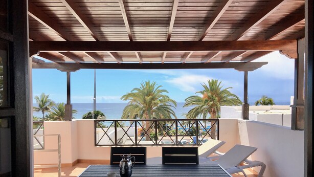 A balcony with a wooden ceiling, a table with a cup and a pitcher, and a view of the sea and palm trees.