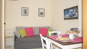 Un comedor con una mesa preparada para dos, un sofá gris con cojines de colores y un televisor empotrado en la pared mostrando un automóvil.