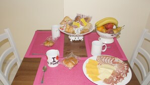 Una mesa de comedor con un mantel individual rosa, sillas blancas y diversos alimentos como pan, frutas, queso y carne.