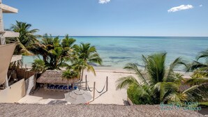 Vista de playa con palmeras, una estructura de techo de paja y un cielo azul despejado.