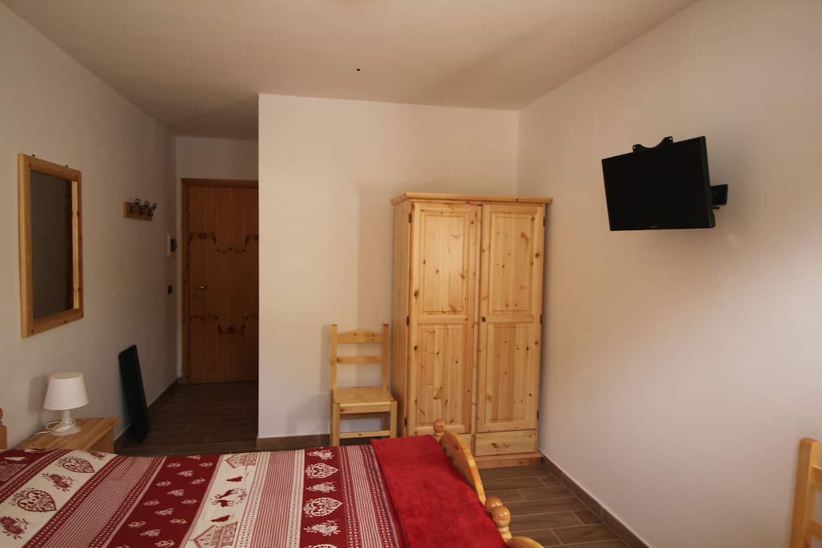 A bedroom with a bed, a wooden wardrobe, a chair, a TV mounted on the wall, and a door leading to another room.