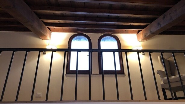 A balcony with wooden beams and a railing, two arched windows with lights, and a chair. - Luxury in the Piazza in Greve In Chiant (Greve in Chianti)