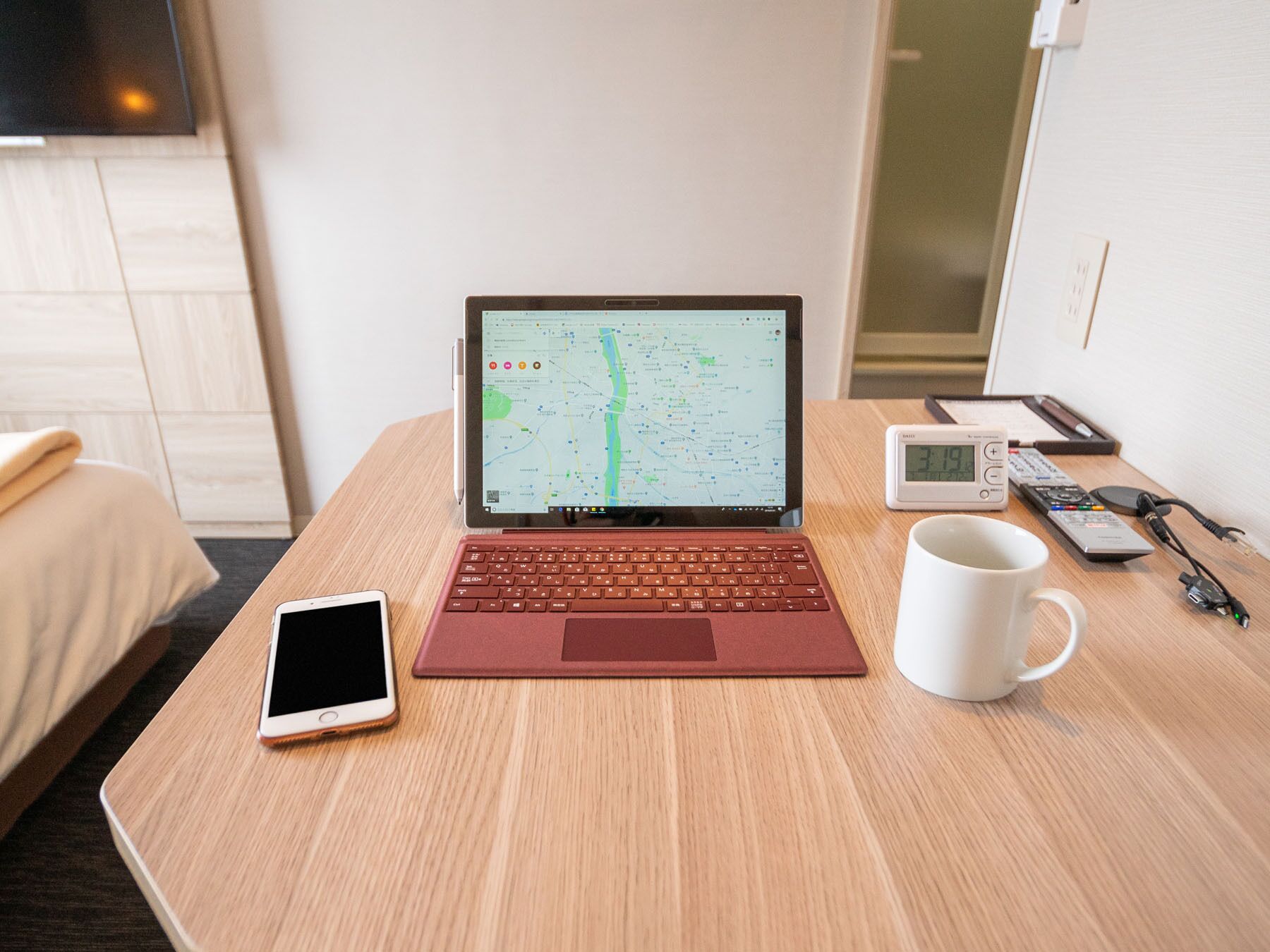 A laptop displaying a map, a smartphone, a mug, a clock, and a remote control on a wooden table in a hotel room.