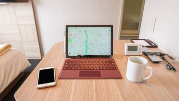 A laptop displaying a map, a smartphone, a mug, a clock, and a remote control on a wooden table in a hotel room.