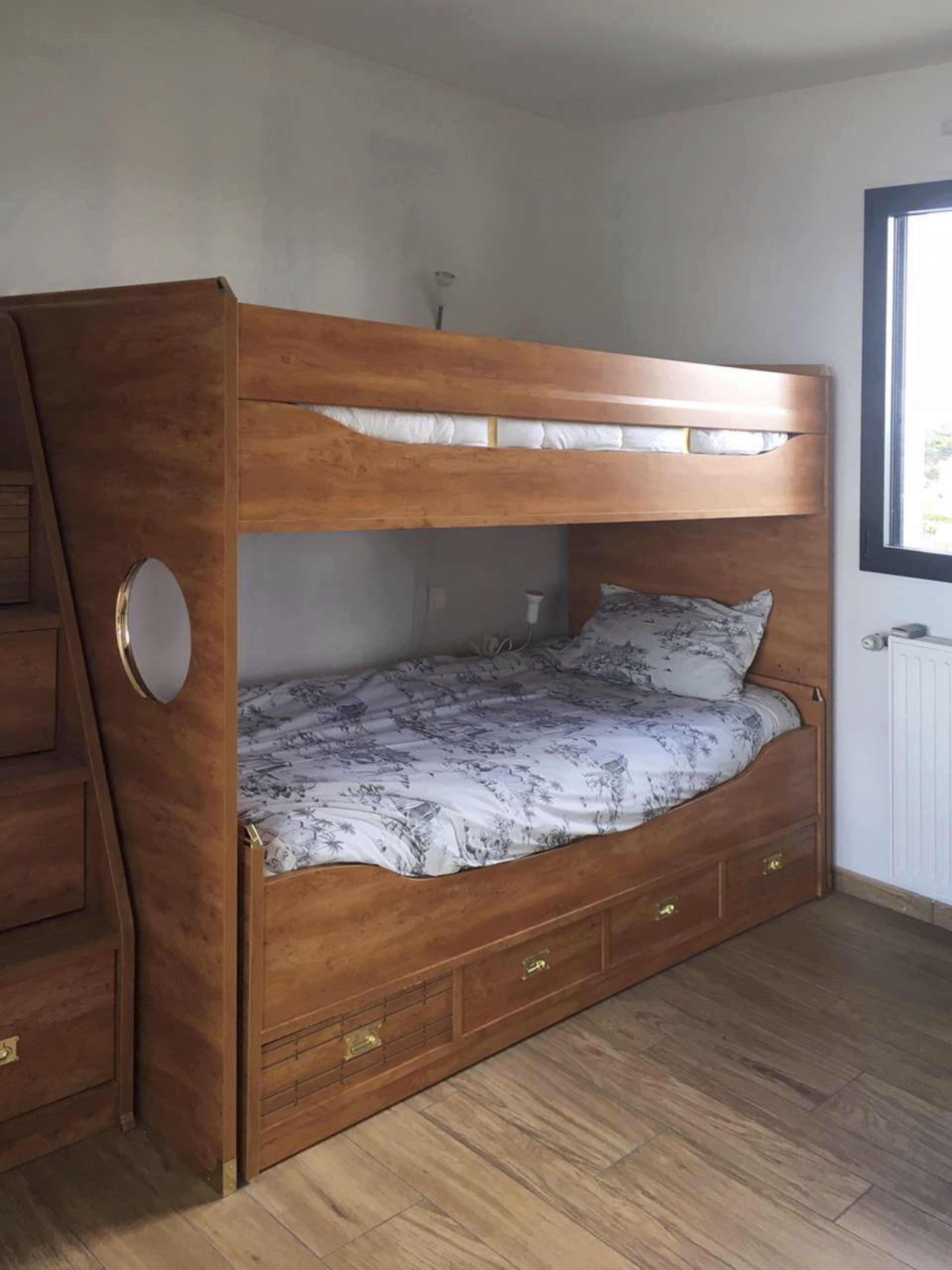 A bunk bed with a wooden frame and drawers, a window with blinds, and a radiator on the wall.