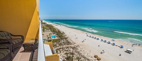 Ein Balkon mit Korbmöbeln, der Blick auf einen Strand mit Sonnenschirmen und Liegestühlen bietet.
