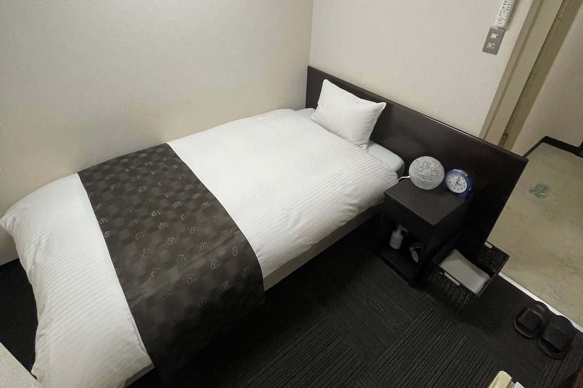 A hotel room with a single bed, a bedside table with a lamp, a clock, and an air conditioning unit mounted on the wall.