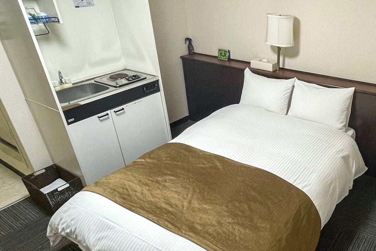 A hotel room with a bed, a nightstand, a lamp, a small table with a clock, a sink, and a cabinet.