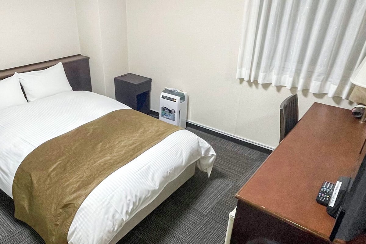 A hotel room with a bed, a desk, a lamp, a trash bin, and a thermostat.