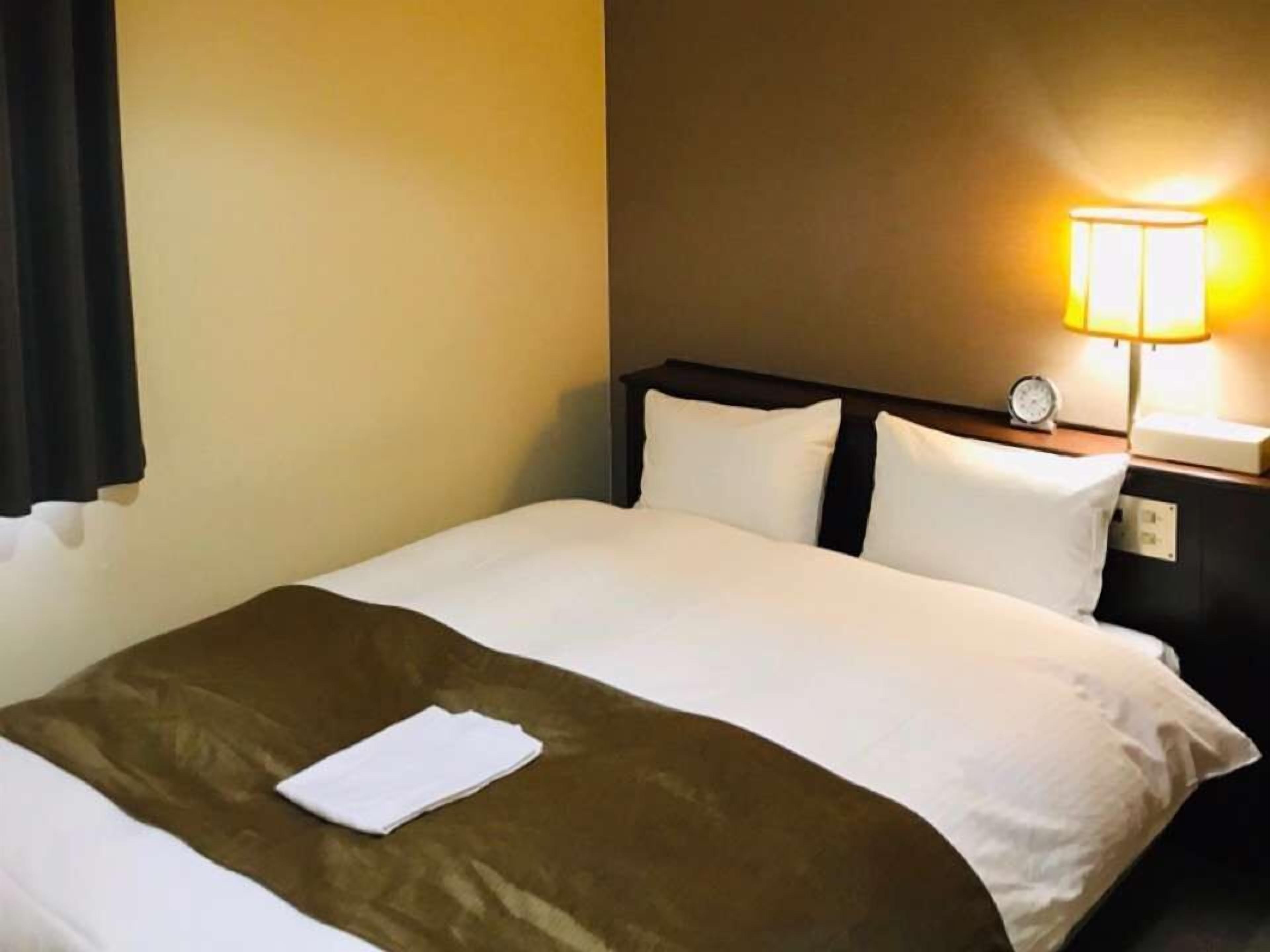 A hotel room with a neatly made bed, a bedside lamp, and a nightstand with a clock.