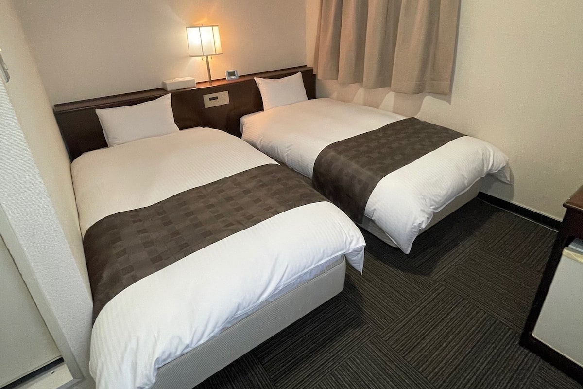 A hotel room with two beds, a bedside table with a lamp, a window with curtains, and a carpeted floor.