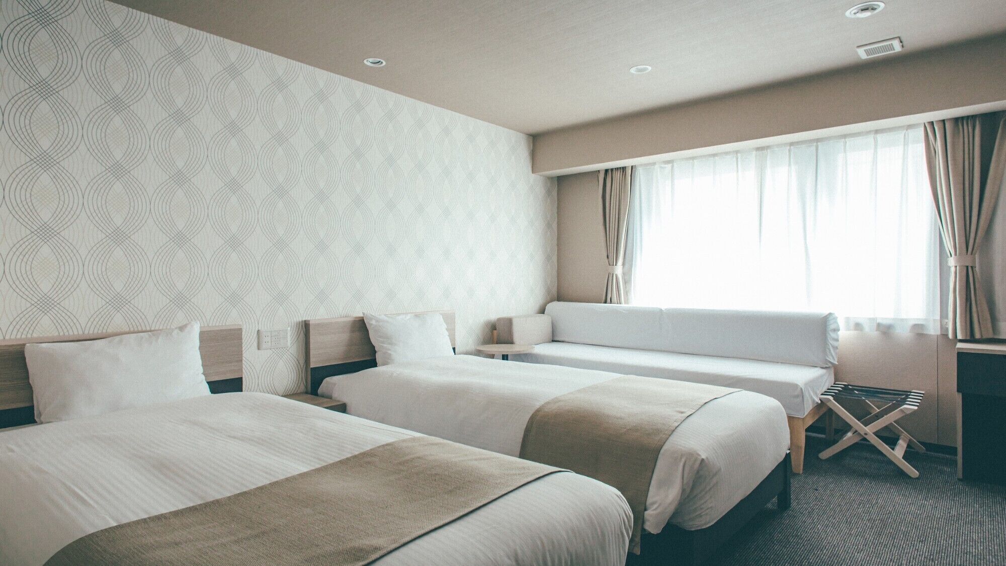 Ein Hotelzimmer mit zwei Betten, einem Schlafsofa, einem kleinen Tisch und einem Fenster mit Vorhängen.