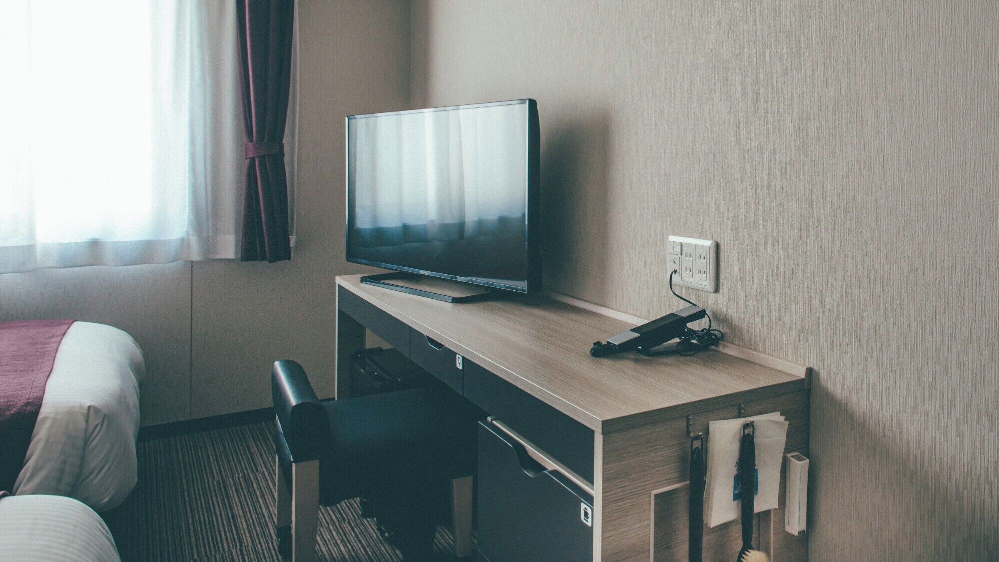 A hotel room with a bed, a desk with a television, a chair, and a power outlet.