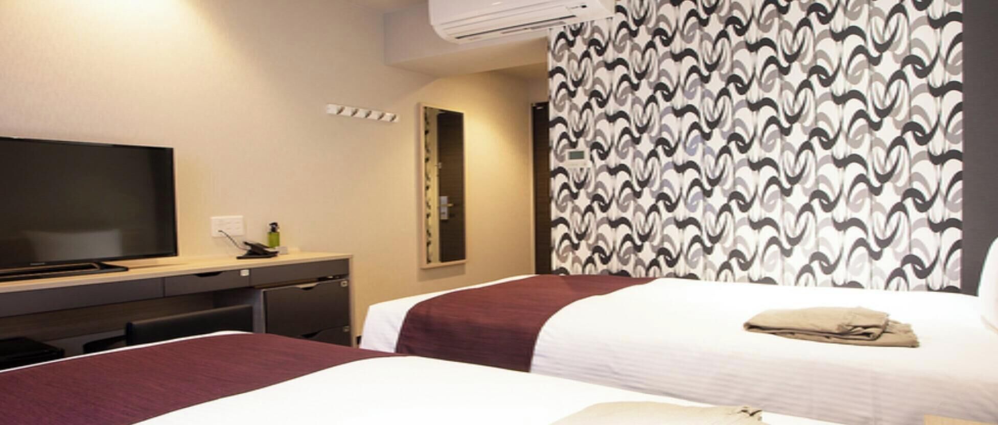 A hotel room with two beds, a desk, a television, and a patterned curtain.