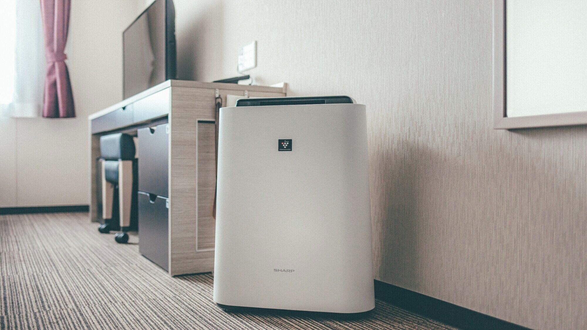 A hotel room with a desk, a chair, a flat-screen TV, and a white air purifier.