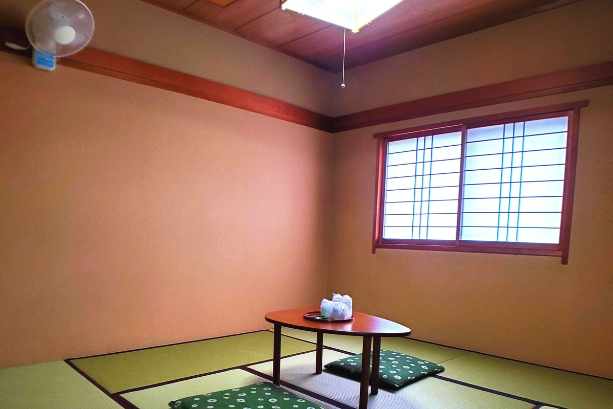Une pièce avec un sol recouvert de tatamis, une petite table et des coussins, un ventilateur de plafond et une fenêtre dotée de panneaux coulissants de type shoji.