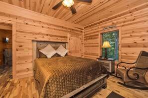 Quarto de cabana de madeira com cama, cadeira, mesa de cabeceira e janela com vista para árvores.