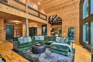 A spacious log cabin interior with a large green sectional sofa, a round ottoman, and a dining area with a table and chairs. - 5 Acres to Roam | Stocked Game Room + Wet Bar  | Our Place to Paws (Lansing)