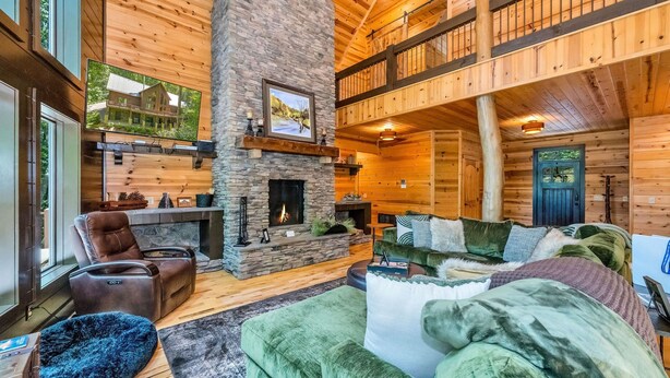 A cozy log cabin interior with a stone fireplace, plush seating, and a balcony overlooking the landscape.