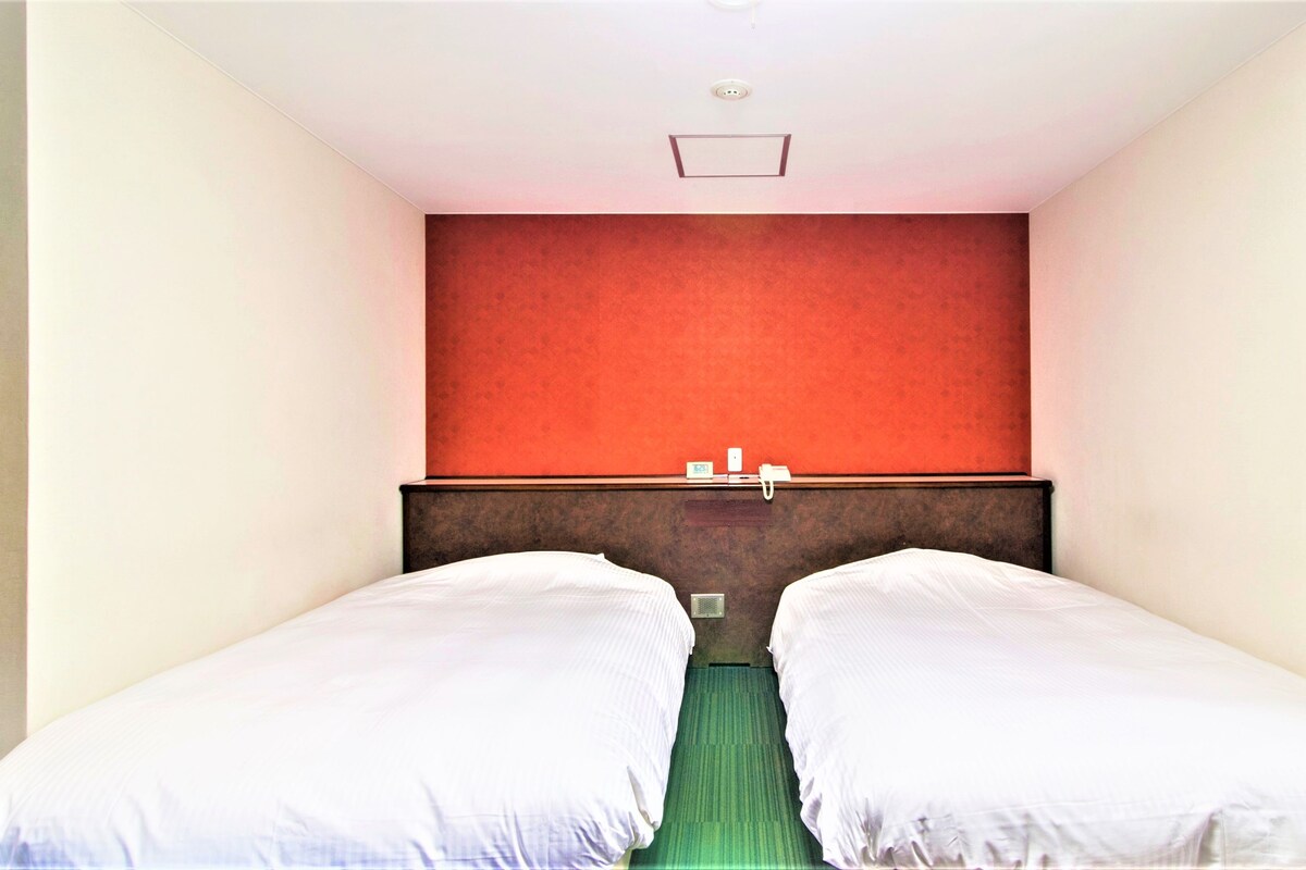 A hotel room with two single beds, a red accent wall, a wooden headboard, a telephone, and a window with curtains.