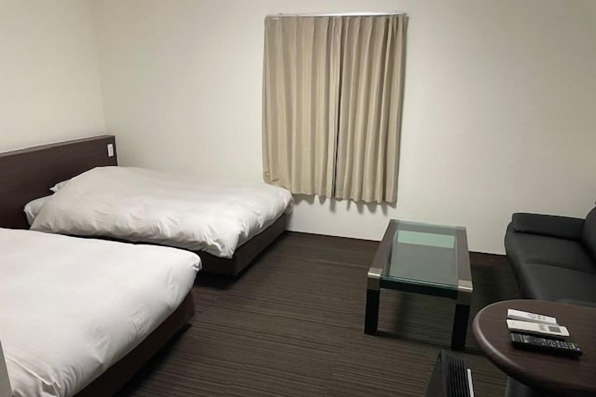 A hotel room with two single beds, a glass-top coffee table, a sofa, and a window with curtains.