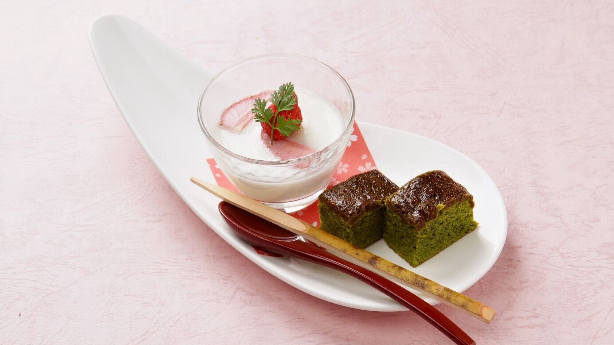 Postre que consisteix en un got de líquid blanc, una guarnició de fragaria, dues porcions de pastís verd i una cullera vermella sobre un plat blanc.