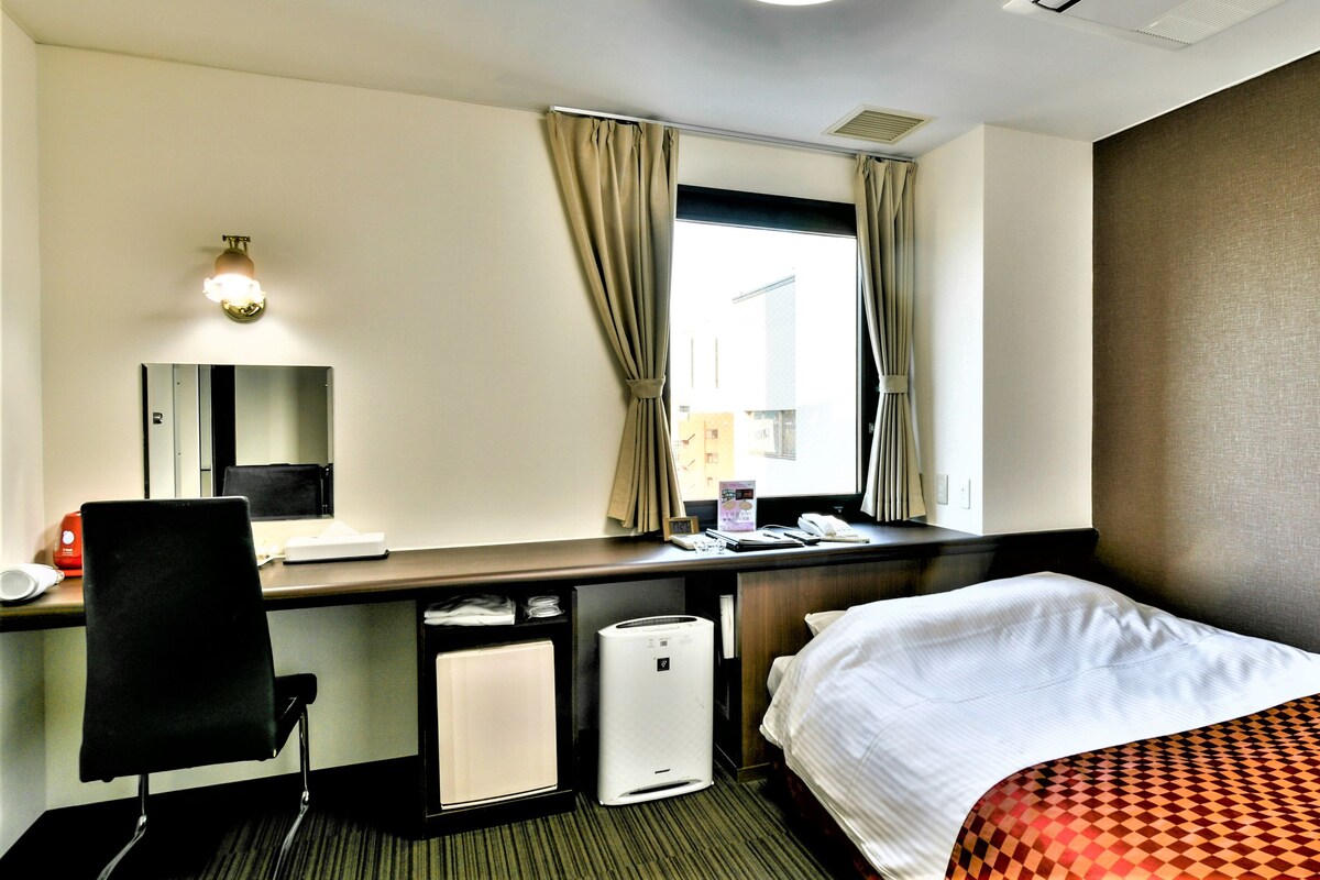 A hotel room with a bed, a desk with a chair, a small heater, a mirror, and a window with curtains.