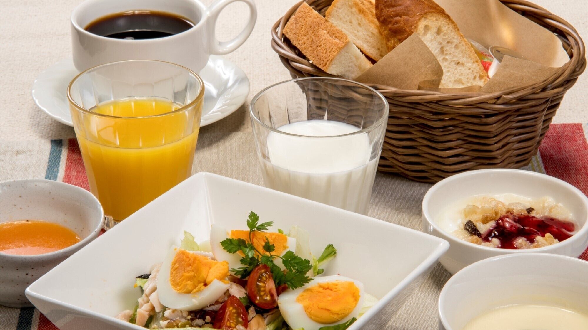 Une table mise pour le petit-déjeuner avec une tasse de café, un verre de jus d’orange, un verre de lait, un panier de pain et divers plats, notamment des œufs, une salade et de la compote de fruits.